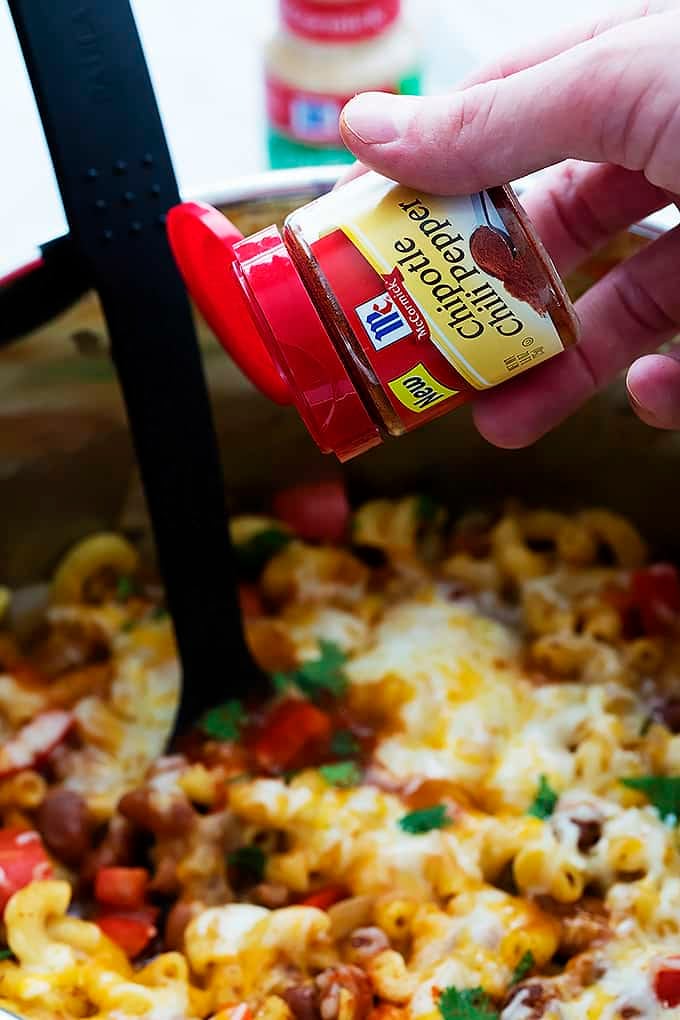 a hand sprinkling come chipotle chili powder on top of cheesy chili mac in a cooking pot.