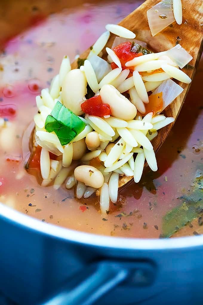 close up of Italian white bean & orzo soup on a wooden spoon above more soup in a cooking pot.