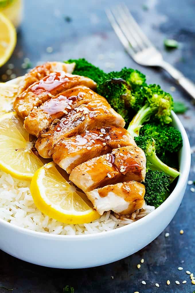 lemon teriyaki chicken with lemon slice and broccoli on rice in a bowl with a fork and more lemon slices in the background.