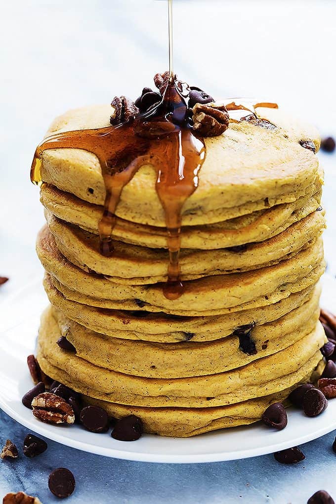 a stack of pumpkin chocolate chip pancakes on a plate topped with nuts and chocolate chips with syrup being poured on top.