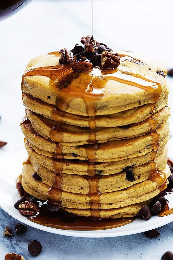 a stack of pumpkin chocolate chip pancakes topped with chocolate chips and nuts with syrup being poured on top.