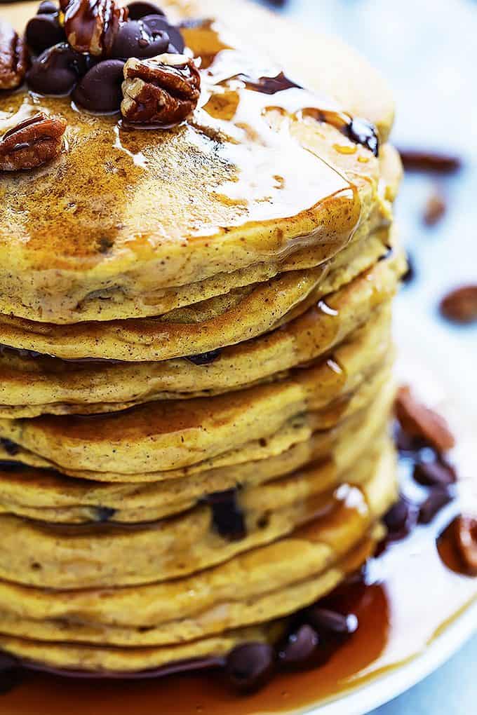 close up of a stack of pumpkin chocolate chip pancakes on a plate topped with chocolate chips, nuts and syrup.