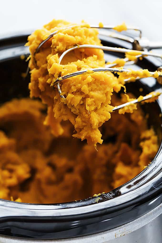 close up of a hand mixer whisks with slow cooker mashed sweet potatoes on them being held above a slow cooker of more sweet potatoes.