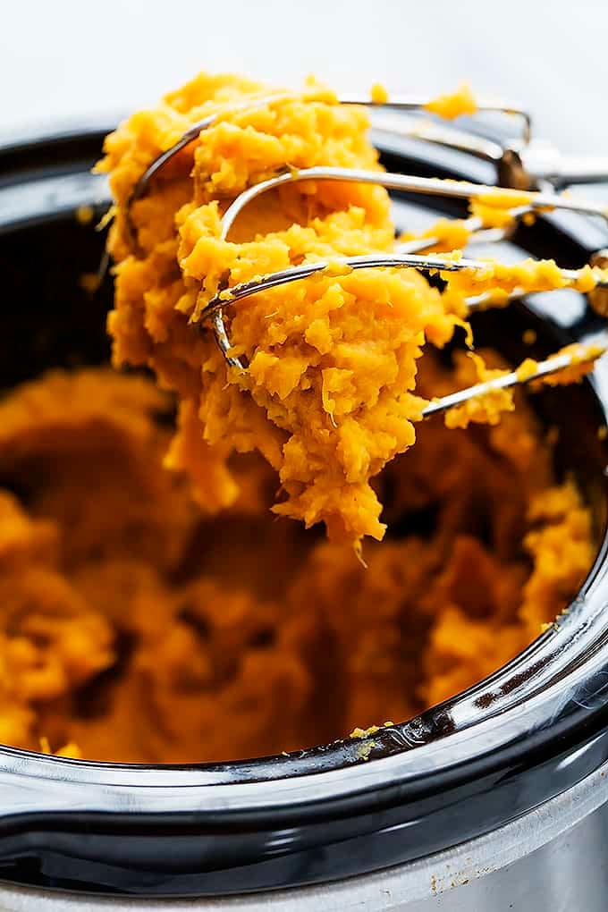 close up of a hand mixer whisks with slow cooker mashed sweet potatoes on them being held above a slow cooker of more sweet potatoes.