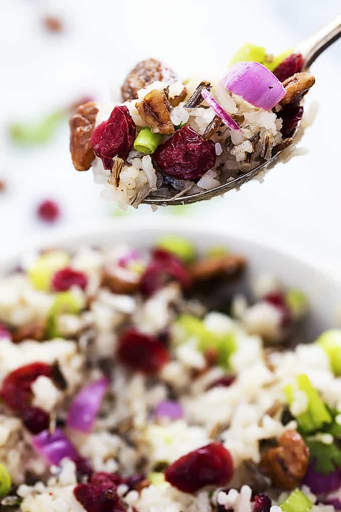 a spoon with cranberry pecan wild rice salad on top being held above a bowl of salad.