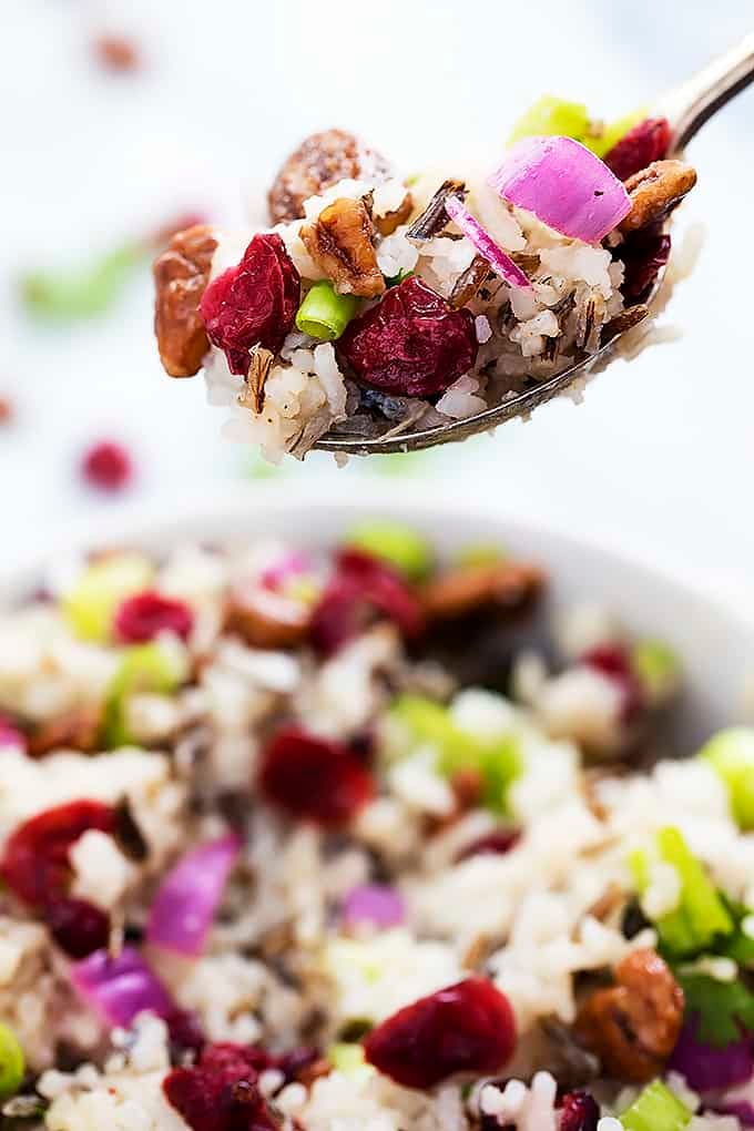 a spoon with cranberry pecan wild rice salad on top being held above a bowl of salad.