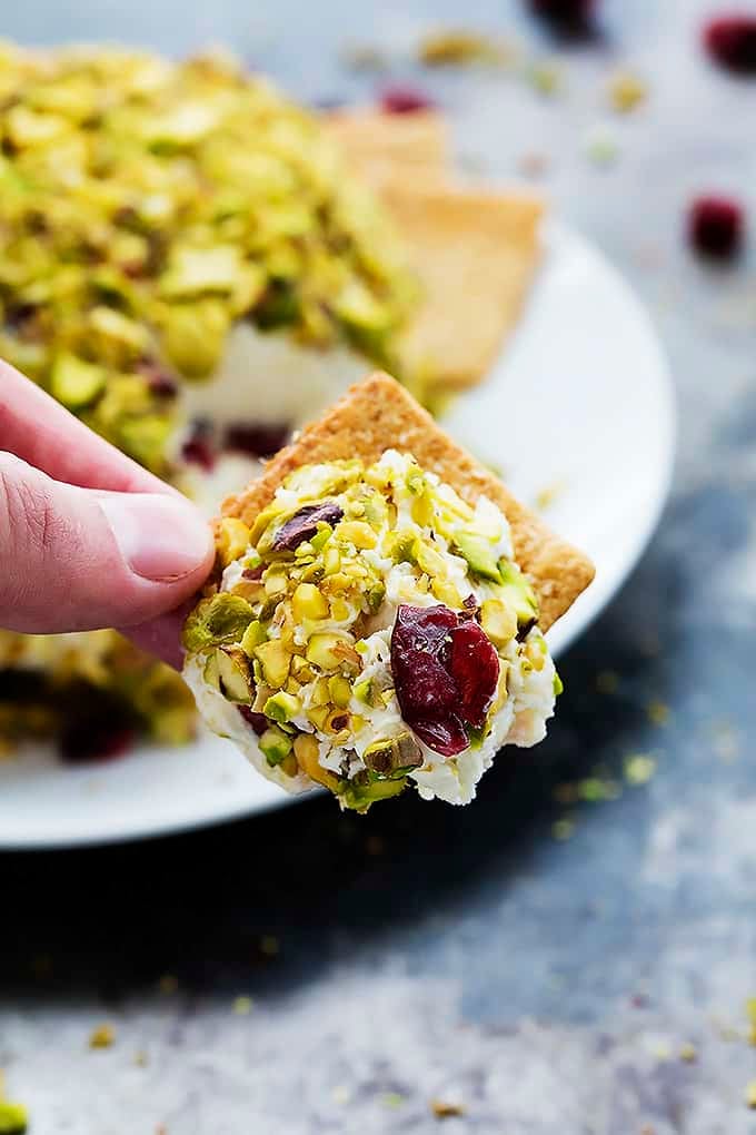 a hand holding up a cracker with some cranberry pistachio cheeseball on top with the cheeseball on a plate in the background.
