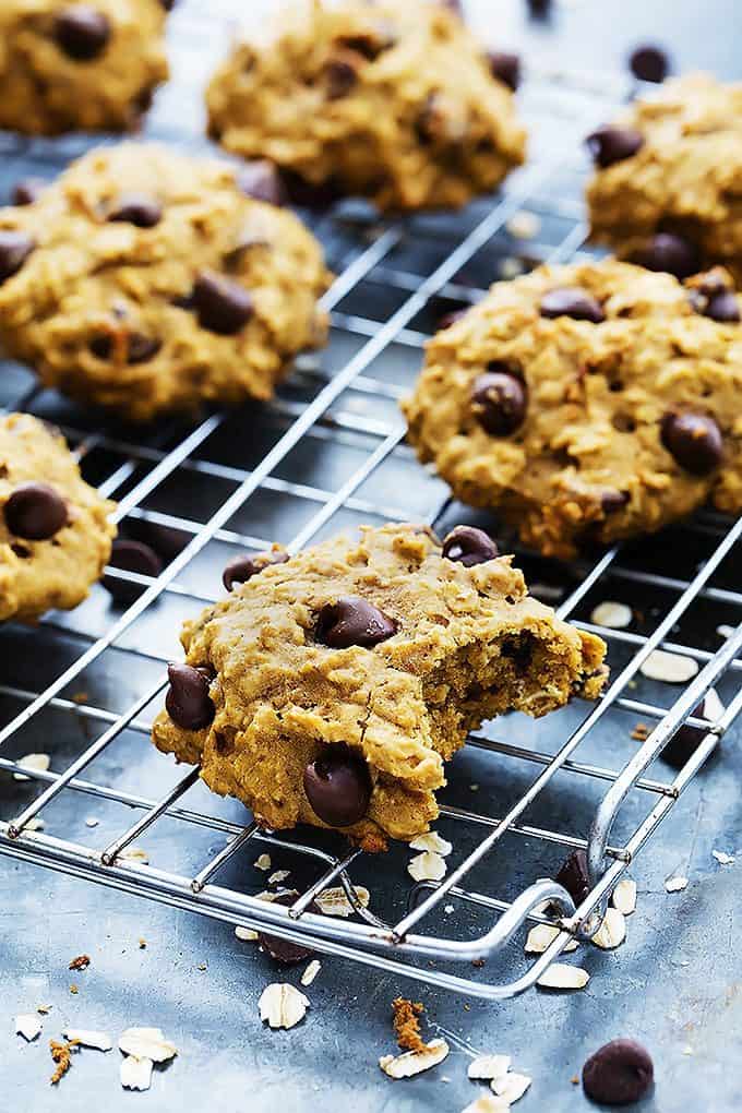 a pumpkin oat chocolate chip cookie with a bite missing with more cookies in the background all on a cooling rack.