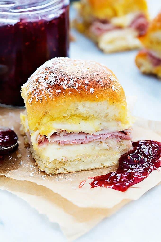 a baked Monte Cristo party slider with a jar of raspberry preserves and more sliders in the background.