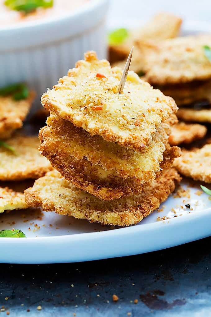 toasted ravioli on a toothpick with more ravioli + cheesy marinara sauce all on a plate.