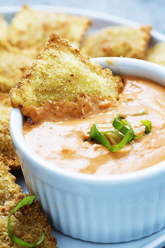 close up of a toasted ravioli dipped in cheesy marinara sauce with more ravioli on the side all on a plate.