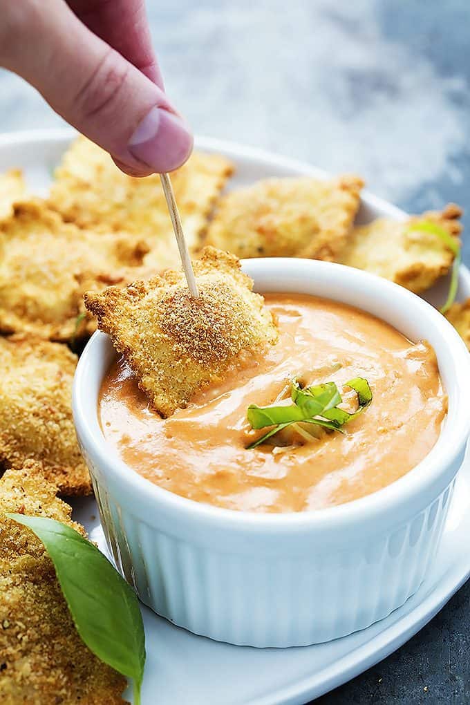 a hand dipping a toasted ravioli in a bowl of cheesy marinara sauce with more ravioli on the side all on a plate.