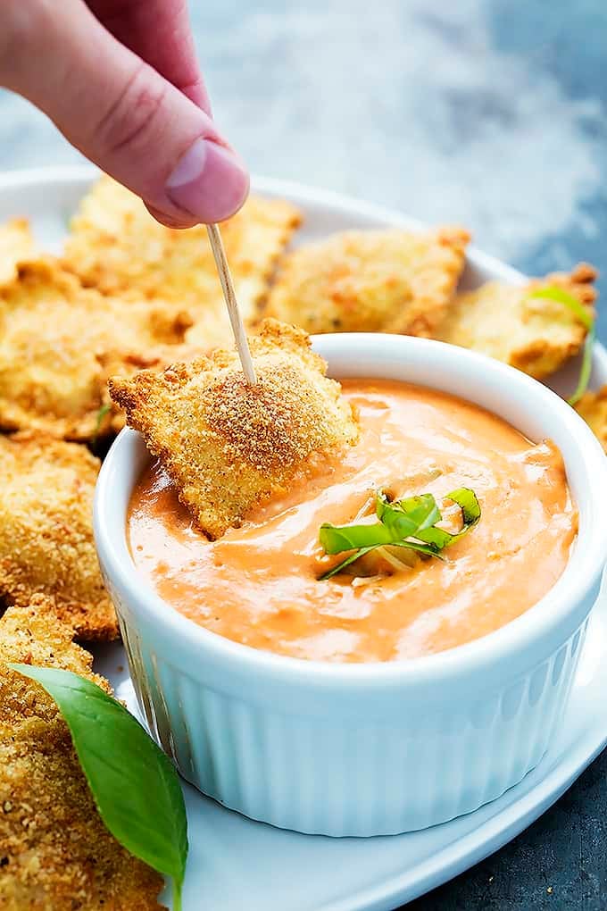 a hand dipping a toasted ravioli in a bowl of cheesy marinara sauce with more ravioli on the side all on a plate.