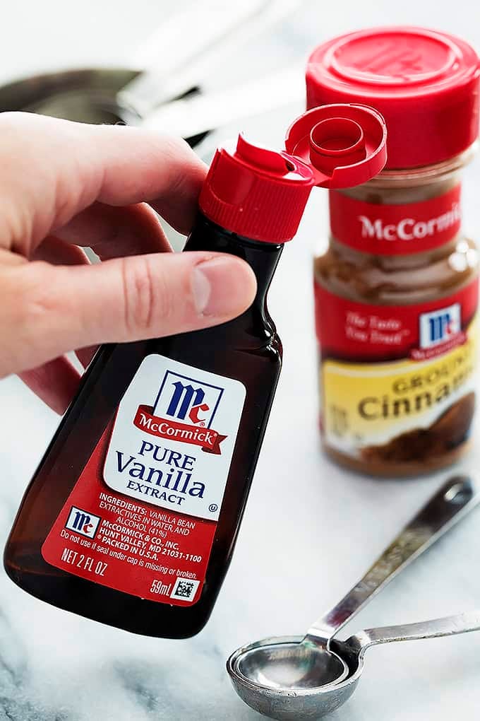 a hand holding a bottle of pure vanilla extract with serving spoons and a container of ground cinnamon in the background.