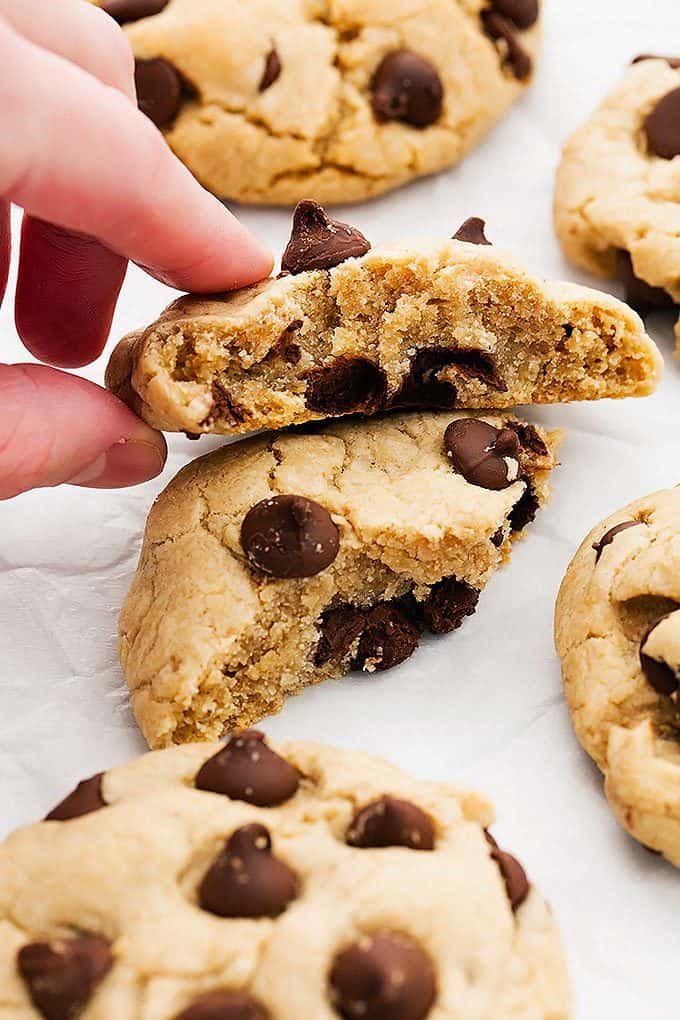a coconut oil cream cheese chocolate chip cookie broken in half with one half laying on top of the other and a hand grabbing the top half with more cookies around it.