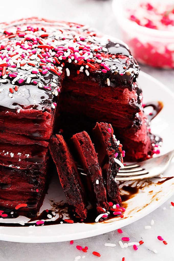 a stack of Nutella stuffed red velvet pancakes topped with sprinkles and chocolate syrup with a bite taken out sitting on a fork all on a plate.