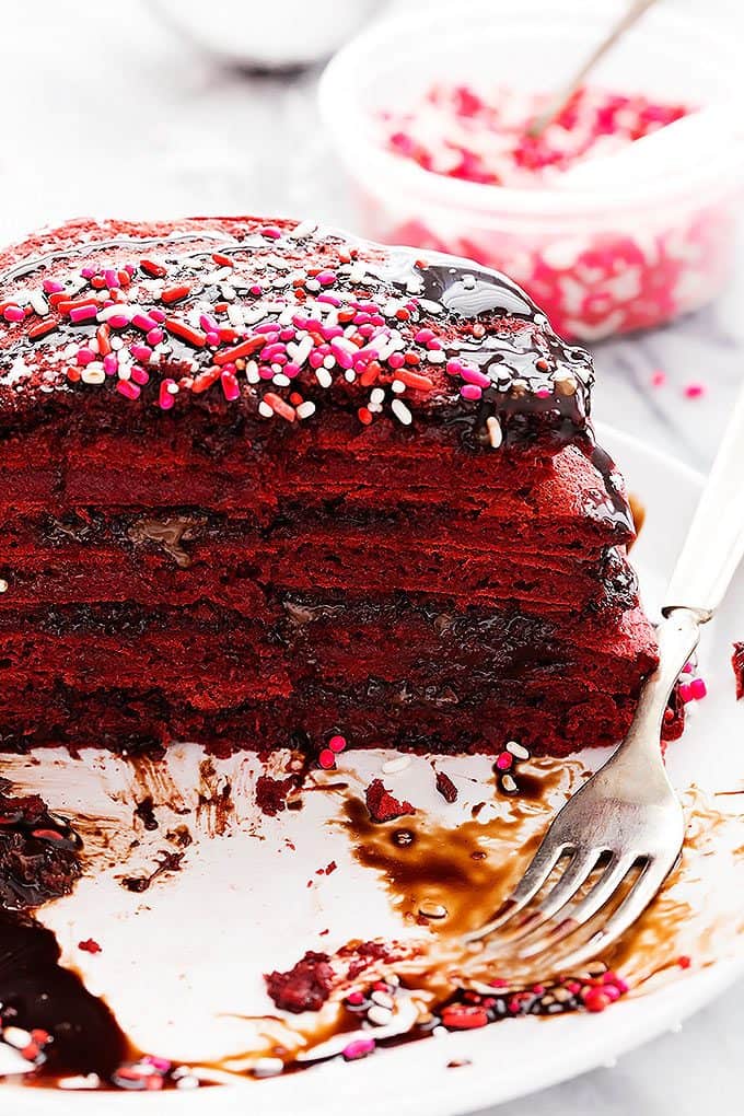 half of a stack of Nutella stuffed red velvet pancakes topped with sprinkles and chocolate syrup with a fork on the side all on a plate.
