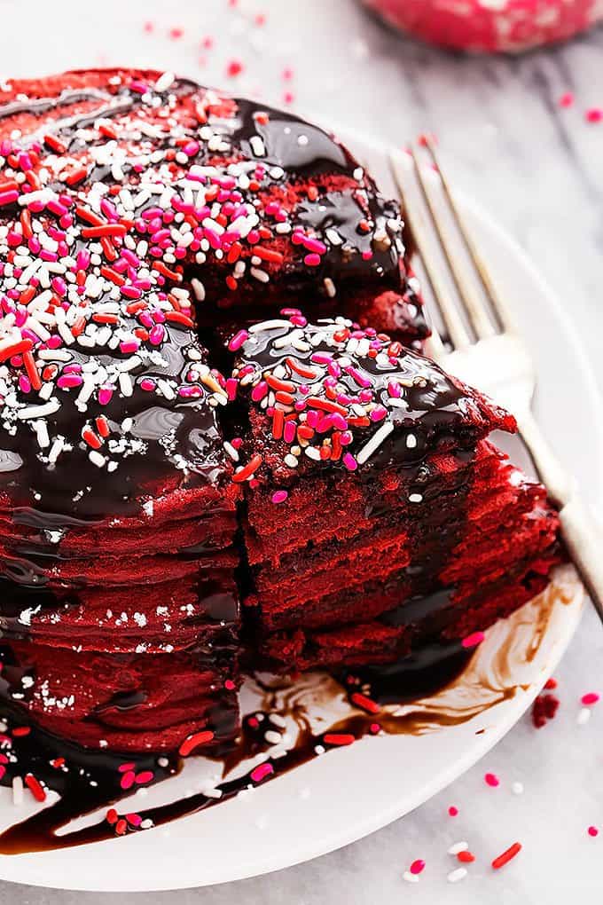 a stack of Nutella stuffed red velvet pancakes topped with chocolate syrup and sprinkles with a bite separated from the rest of the pancakes with a fork on the side all on a plate.