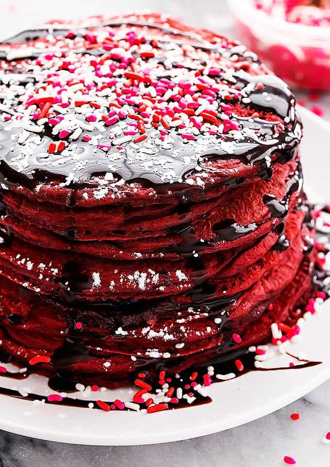 a stack of Nutella stuffed red velvet pancakes on a plate topped with chocolate syrup and sprinkles.