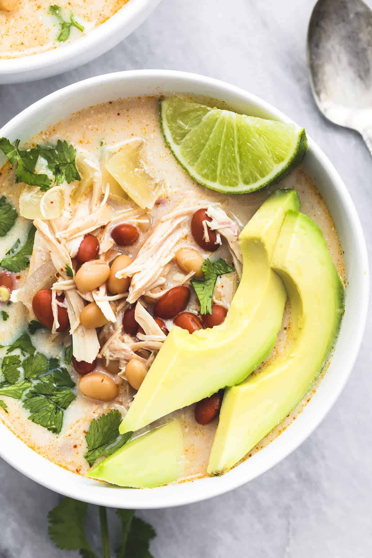 top view of slow cooker cream cheese chicken chili topped with avocado slices and a lime slices with a spoon and another bowl of chili on the side.