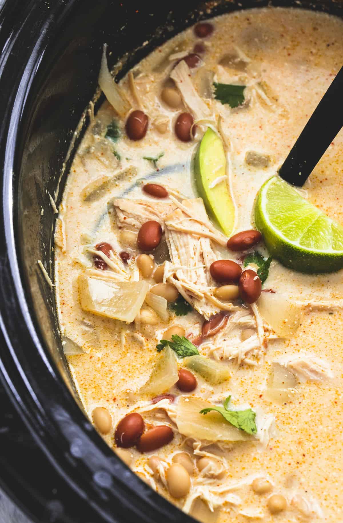 close up of slow cooker cream cheese chicken chili with lime slices and a serving spoon in a slow cooker.