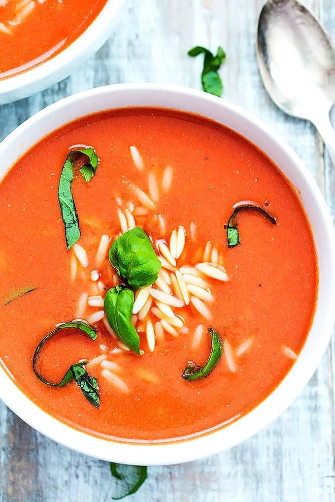 close up top view of tomato basil orzo soup in a bowl with another bowl of soup and a spoon on the side.