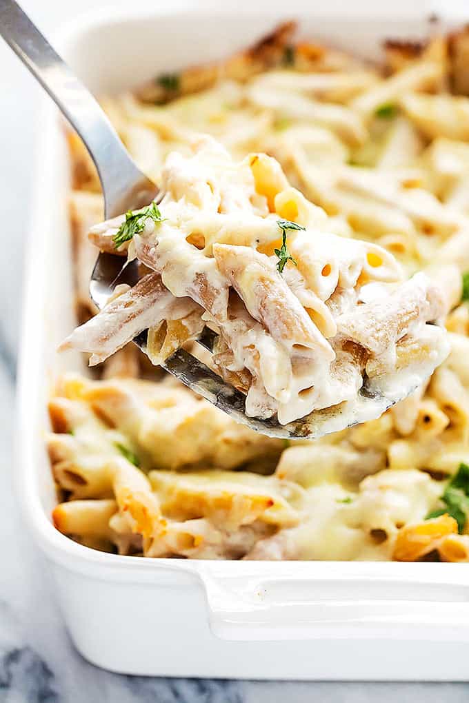 a spatula with chicken alfredo baked penne on it being held up in front of a baking pan of more pasta.