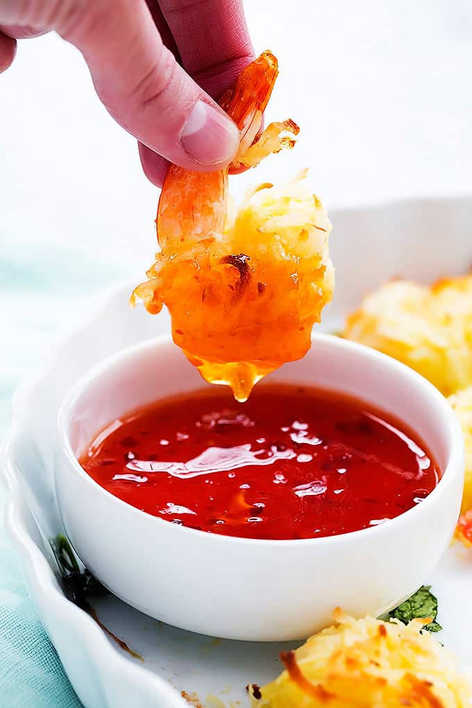  a hand picking up a baked coconut shrimp just dipped in a bowl of sweet and sour sauce.