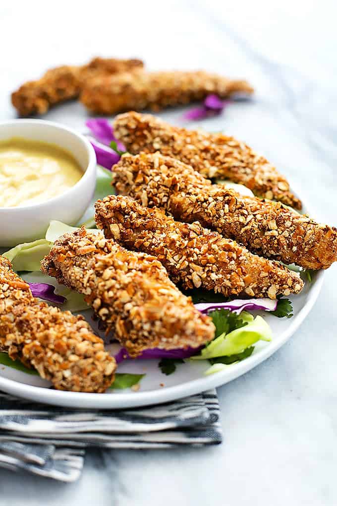 baked pretzel crusted chicken tenders and dipping sauce on a plate more with tenders in the background.