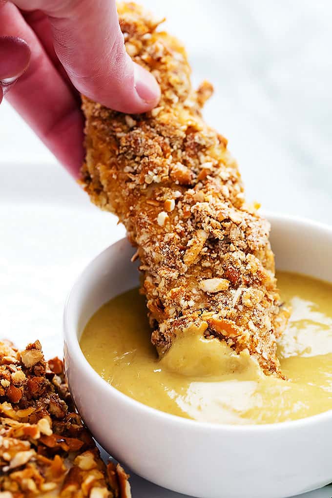 close up of a hand dipping a baked pretzel crusted chicken tender in dipping sauce.