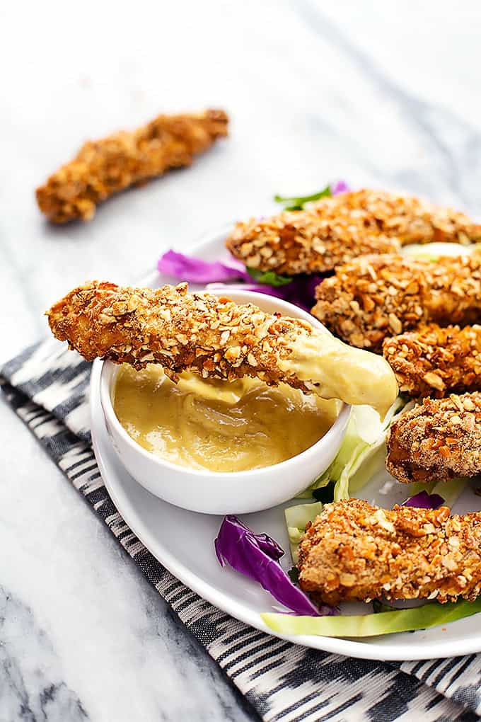 a baked pretzel crusted chicken tender dipped in sauce sitting on top of a bowl of dipping sauce with more tenders on the side all on a plate.