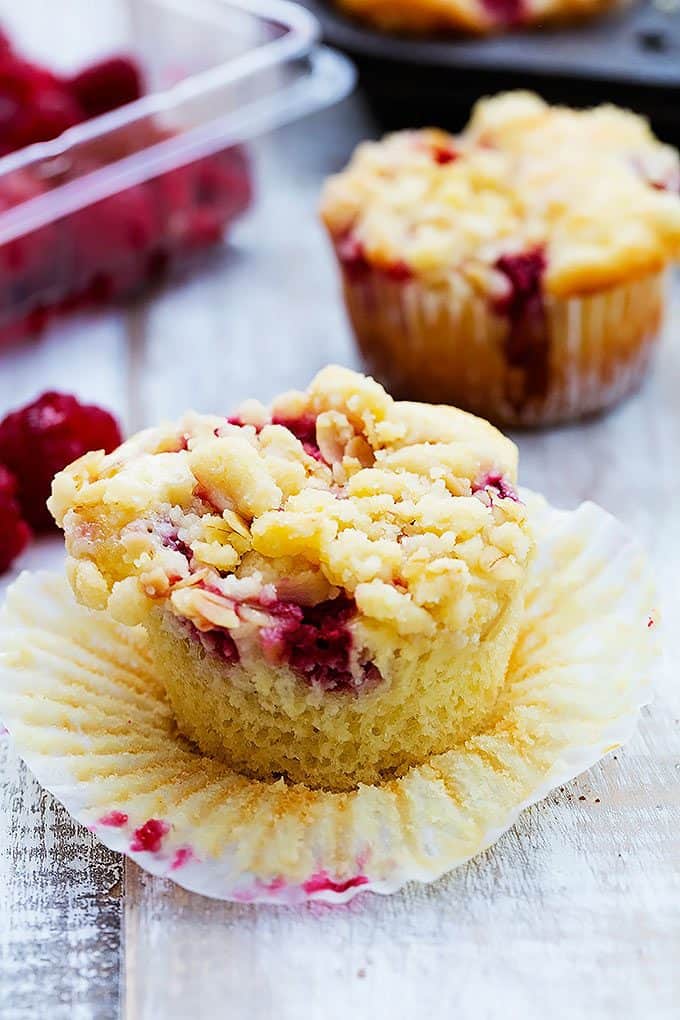 a raspberry crumb muffin with the liner peeled off with raspberries and another muffin in the background.