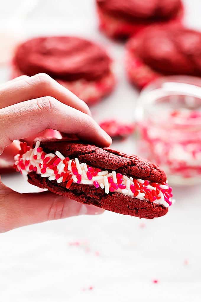 a hand holding up a red velvet sandwich cookie with a jar of sprinkles and more cookies in the background.