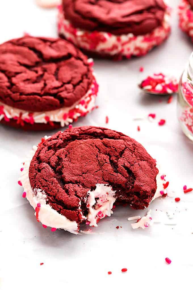 a red velvet sandwich cookies with a bite missing with more cookies and a spoon and jar both full of sprinkles in the background.