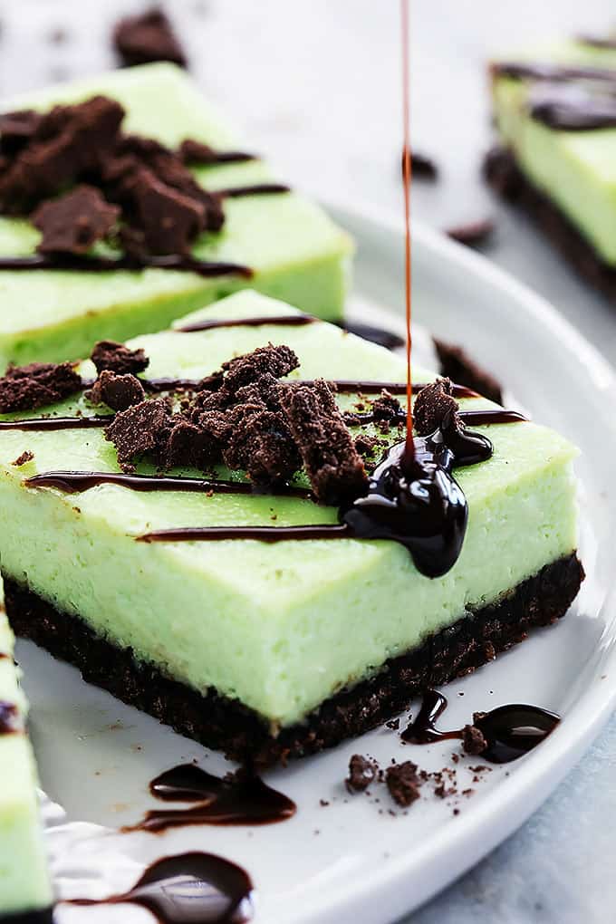 a thin mint cheesecake bar with chocolate sauce being poured on top with more bars around it on a plate.