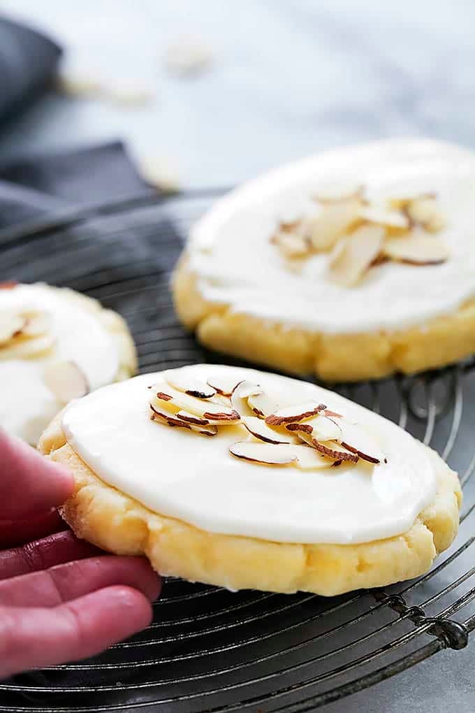 close up of a hand taking a almond sugar cookie from a round cooling rack with more cookies on the side.