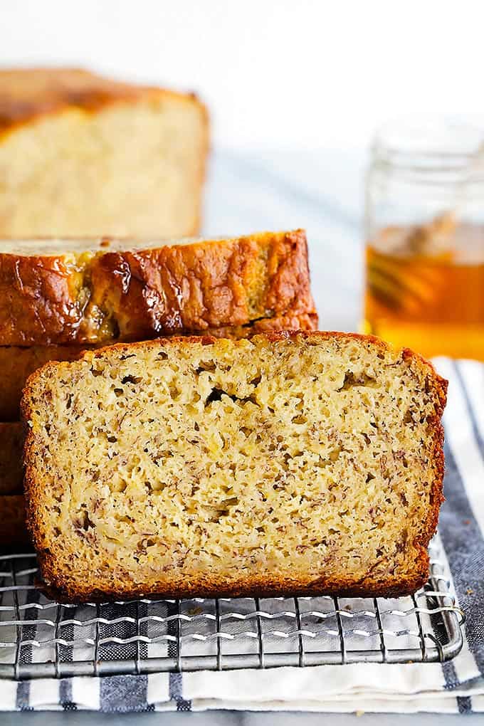 a slice of brown butter banana bread leaning on a stack of more slices of bread on a cooling rack with the rest of the loaf and a jar of honey in the background.