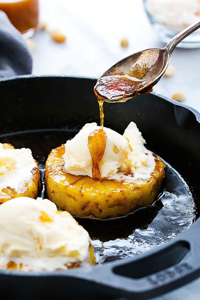 close up of caramelized pineapple sundaes in a cast iron with a spoon pouring the pan sauce on one pineapple sundae.