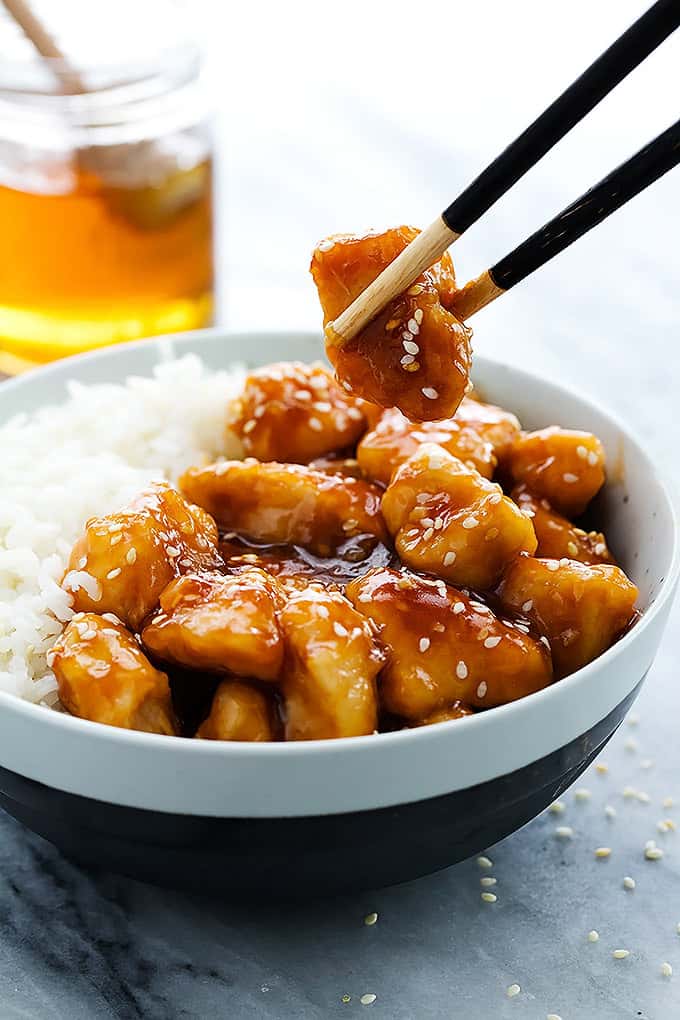 a pair of chopsticks holding a piece of slow cooker honey sesame chicken above a bowl of chicken and rice with a jar of honey with a honey dipper inside in the background.