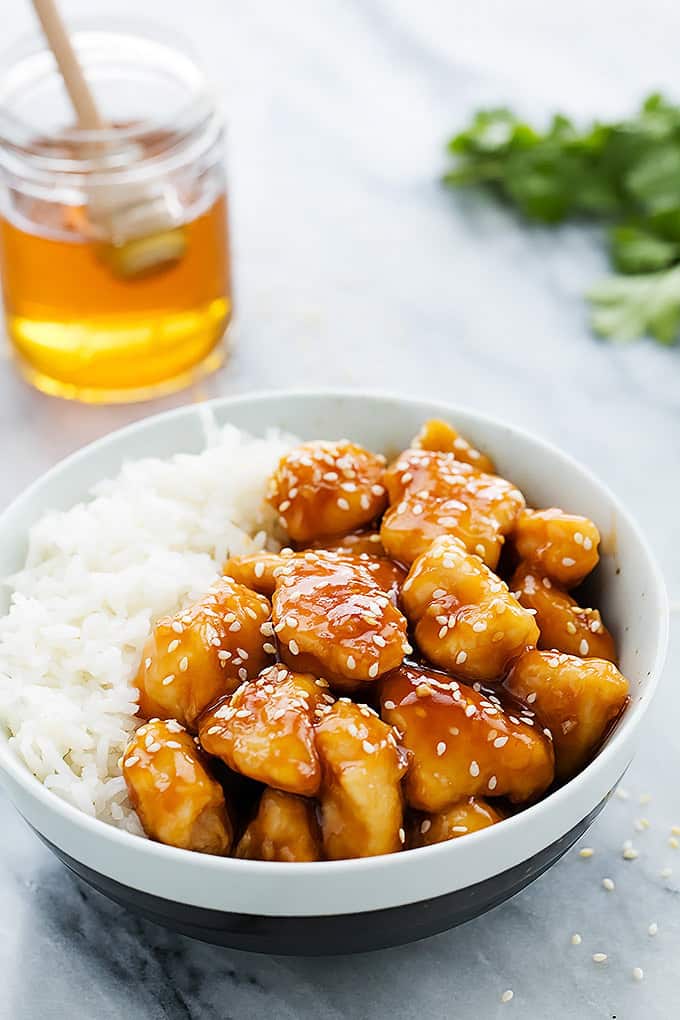 slow cooker honey sesame chicken with rice in a bowl with a jar of honey with a honey dipper in it in the background.