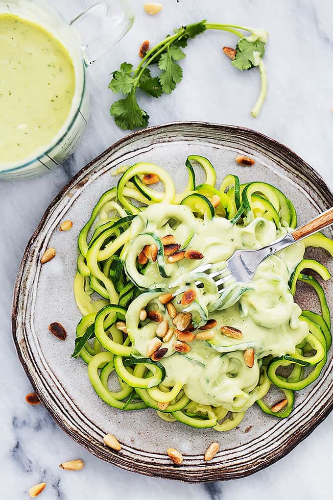 top view of zoodles (zucchini noodles) with cilantro lime avocado sauce topped with toasted pine nuts and a fork on a plate with more sauce on the side.