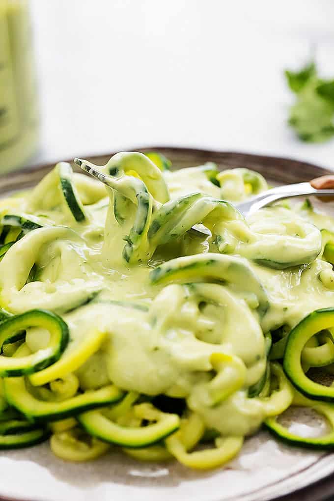 close up of a fork picking up some zoodles (zucchini noodles) with cilantro lime avocado sauce from a plate.