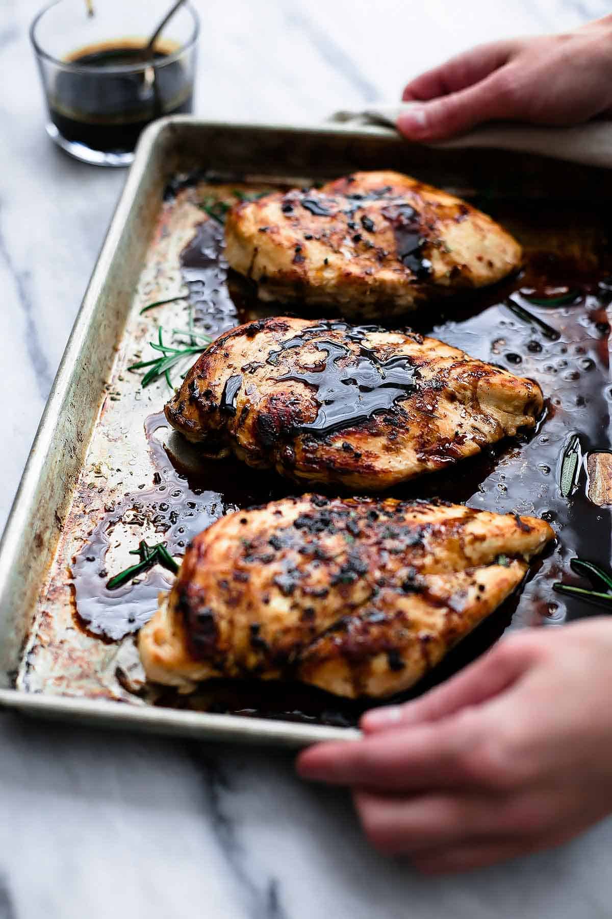 hands holding a baking sheet of baked balsamic chicken with a glass of balsamic vinegar with a spoon in it on the side.