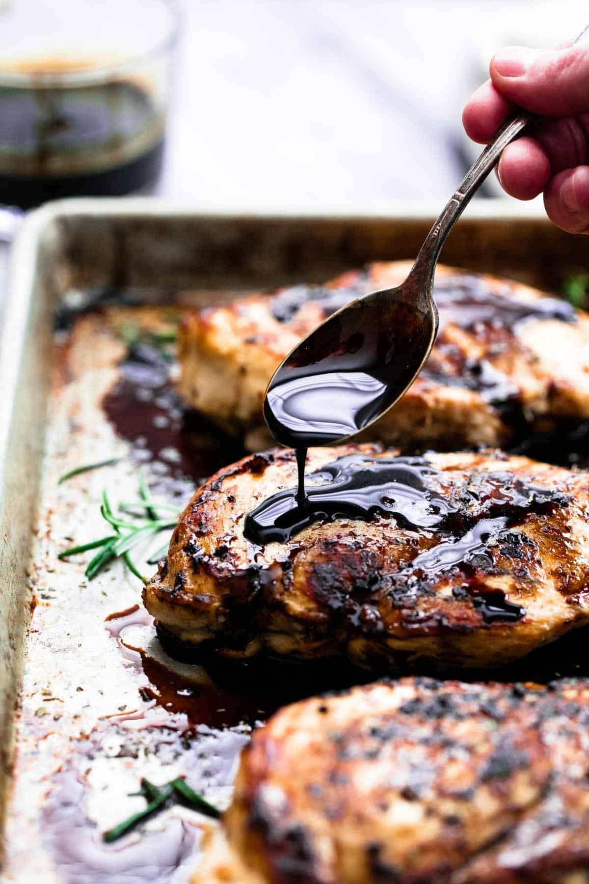 a hand pouring balsamic vinegar on baked balsamic chicken on a baking sheet with a spoon.