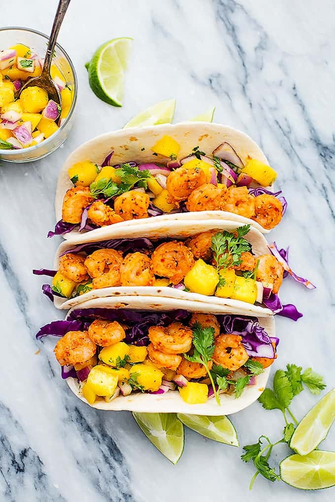 top view of Cajun shrimp tacos with mango salsa with lime slices and a cup of more salsa with a spoon in it on the side.