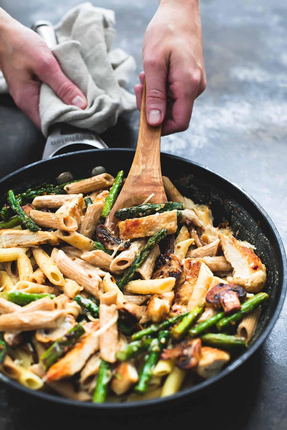 a hand scopping some garlic chicken mushroom and asparagus penne from a skillet with a wooden spoon.