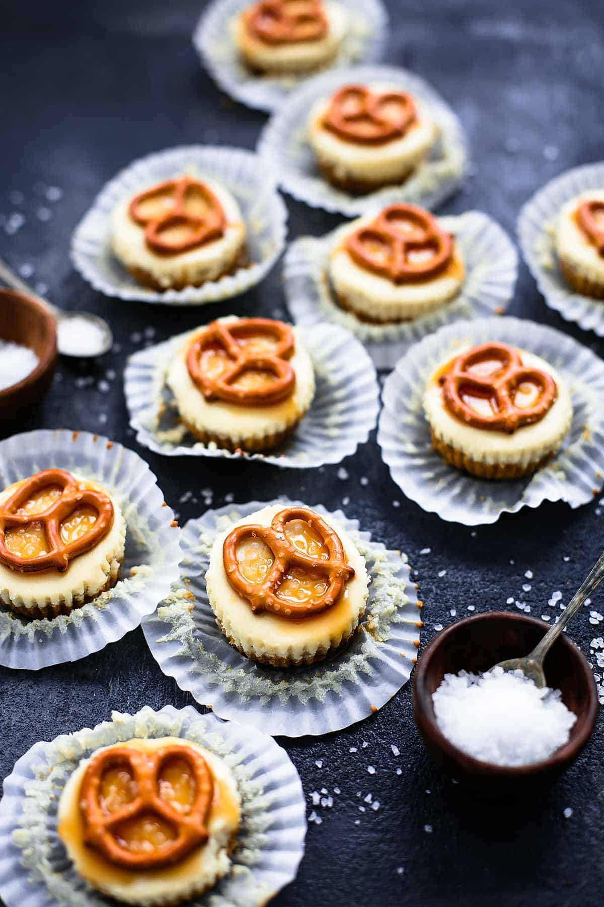 salted caramel pretzel mini cheesecakes with their liners open with small bowls and spoons with salt on top on the side.