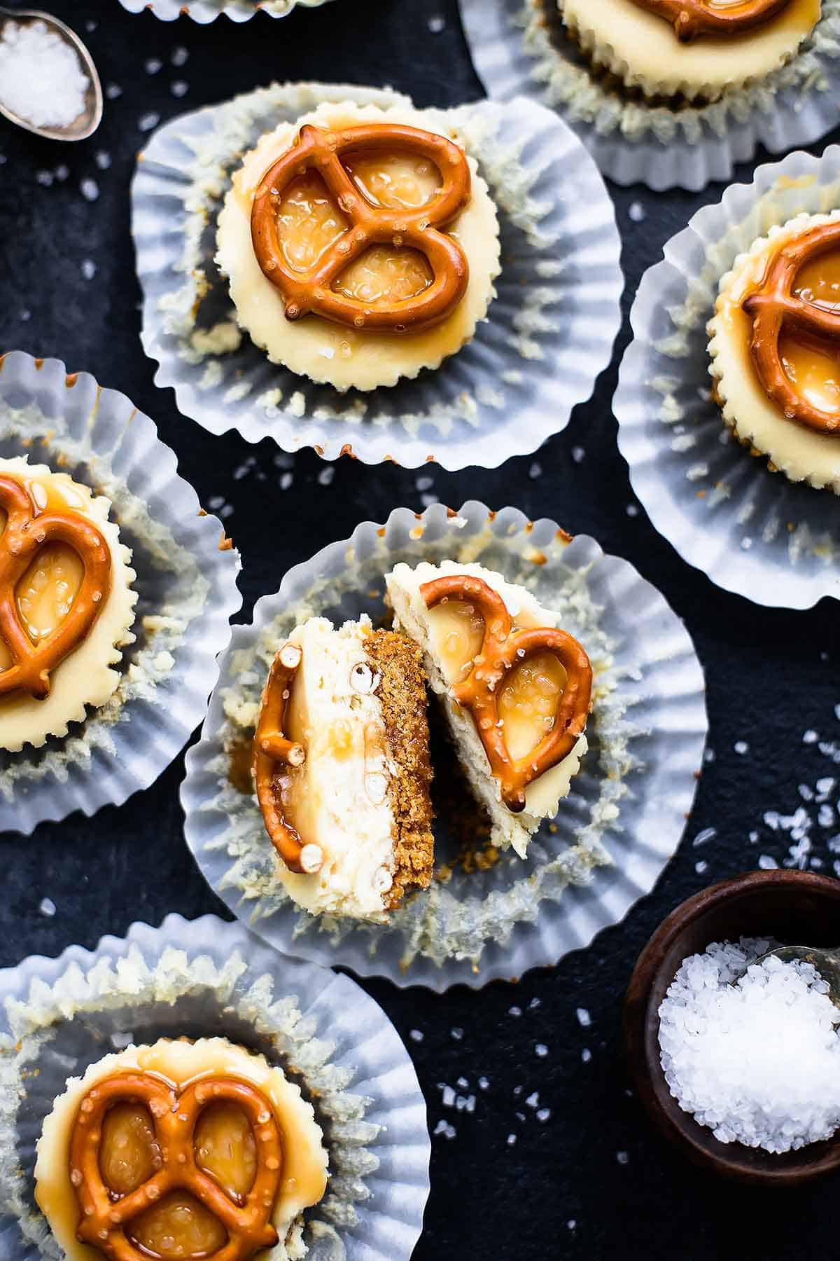 top view of a salted caramel pretzel mini cheesecake cut in half with more cheesecakes around it with their liners opened.