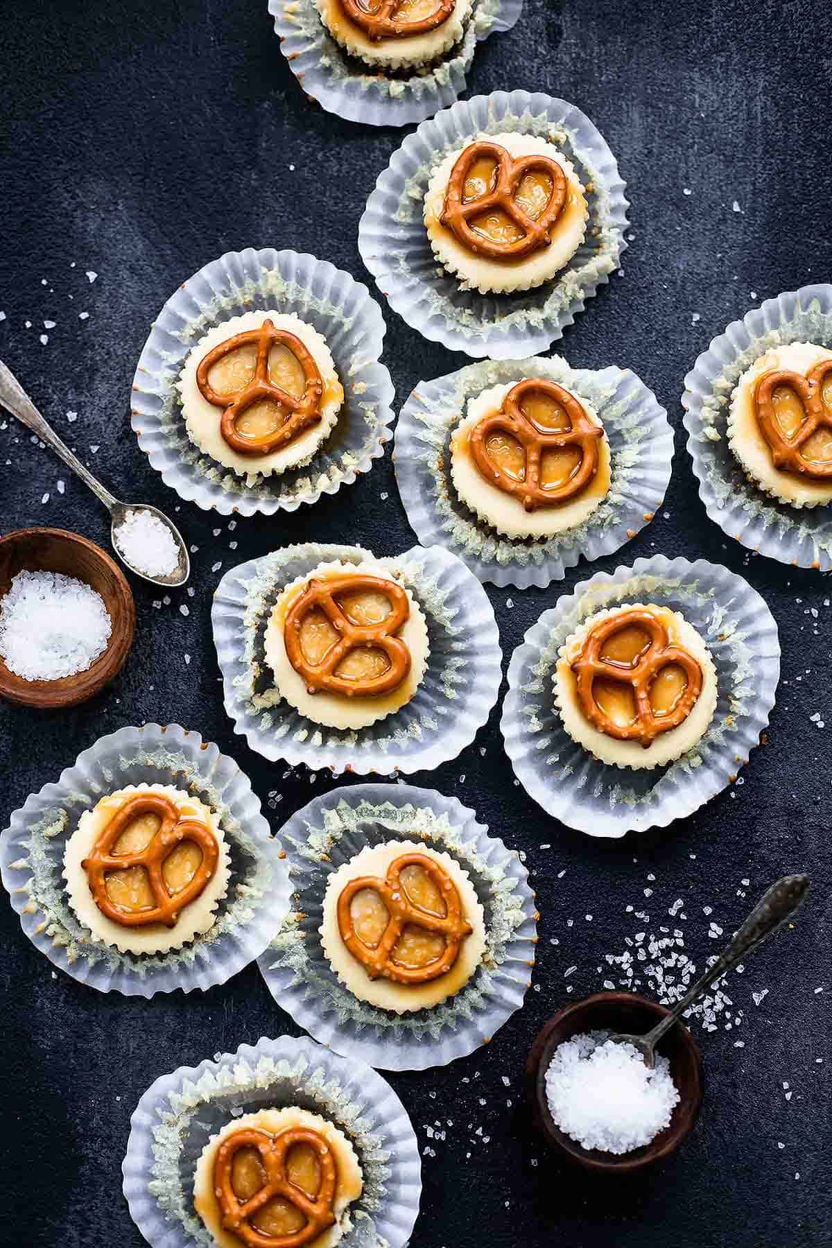 top view of salted caramel pretzel mini cheesecakes with their liners open with salt in small bowls and on spoons on the side.