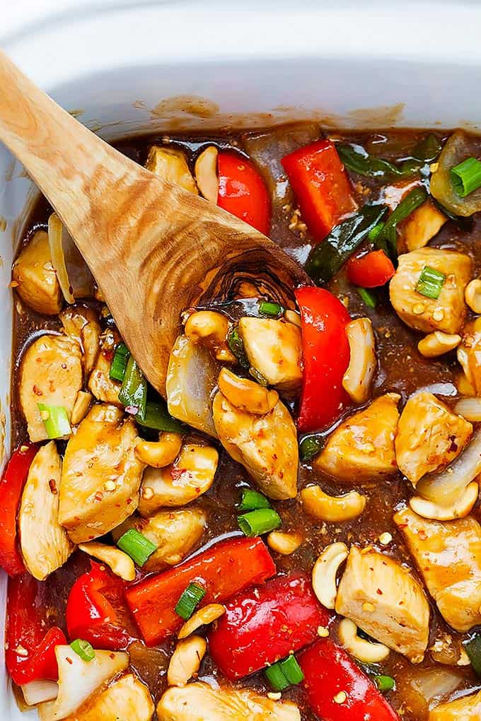 close up of slow cooker spicy cashew chicken and a wooden serving spoon in a serving pan.